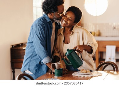 Black couple with afro hair enjoy a romantic breakfast moment in their kitchen. Happy wife leaning in for a loving cheek kiss while pouring filtered coffee at the breakfast table at home. - Powered by Shutterstock