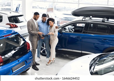 Black Couple Buying New Car Choosing Auto With Manager Standing In Automobile Dealership Center. Copy Space