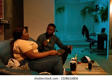 Black Couple Eating Chips And Watching TV On Sofa At Home. Concept Of Domestic Entertainment And Leisure. Idea Of Relationship And Spending Time Together. Man Looking At His Girlfriend. Evening Time