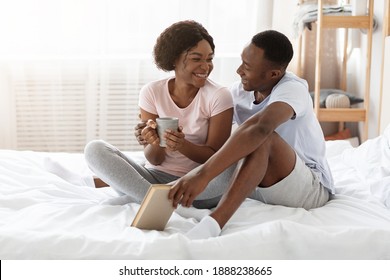 Black Couple In Love Sitting On Bed At Home And Talking, Sharing Thoughts And Dreams. Happy African American Man And Woman In Pajamas Drinking Tea And Reading Book, Spending Weekend Together