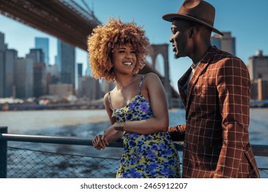 Black couple smiling and talking to each other next to a metal fence, standing on a river promenade with New York City in the background. - Powered by Shutterstock