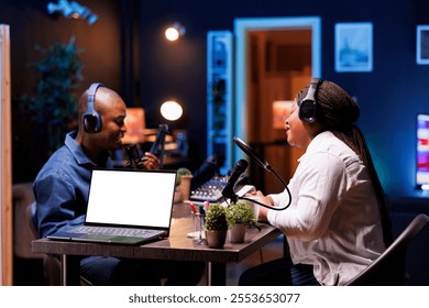 Black couple wearing headphones and talking on microphones while laptop shows isolated white screen. Device on table displays blank chromakey mockup template while two vloggers creates online content. - Powered by Shutterstock