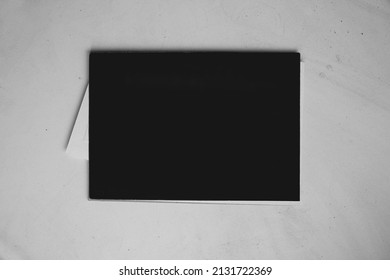 Black Cover Page Of Small Learning Sketchbook With White Small Paper Card Inside, On White Texture Dusty Table Background, For Branding Identity Advertising Mockup.