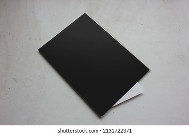 Black Cover Page Of Small Learning Sketchbook With White Small Paper Card Inside, On White Texture Dusty Table Background, For Branding Identity Advertising Mockup.