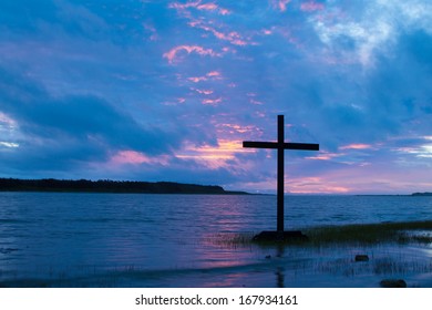 Black cross by water with a  clouded sky as the sunsets.