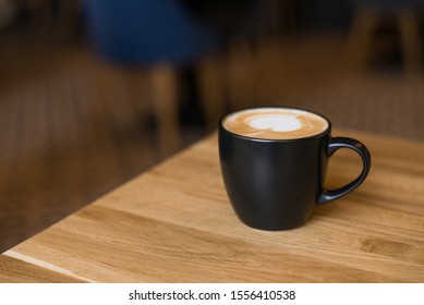 black cup with cappuccino on a wooden table, in the background the coffee shop hall, horizontal banner, the item is located at the bottom right, place for text