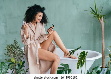 Black curly female wearing bathrobe sits on bathtub choosing between plastic or glass bottle with cosmetic care product. Zero waste concept. Green and conscious lifestyle concept.