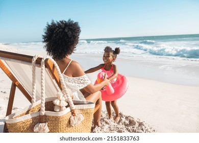 Black cute little girl inside pink float standing on beach with mother while playing together. Rear view of young african american mother sitting on deck chair holding hands of her daughter at seaside - Shutterstock ID 2151833763
