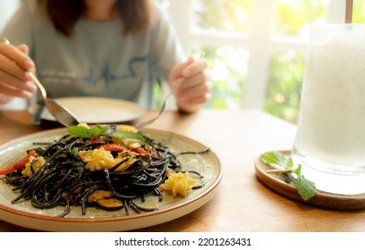 Black Cuttlefish Ink Spaghetti With Shrimp On Plate. Black Pasta With Squid Ink On A Restaurant Table And Blur Woman Eating With Fork And Spoon. Healthy Food. Woman Eating Delicious Black Spaghetti.