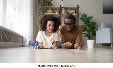 Black dad daughter school age assemble jigsaw puzzle put together pieces lying on warm heated floor in living room. Leisure hobby having fun, logical reasoning ability, develop solving skills concept - Powered by Shutterstock
