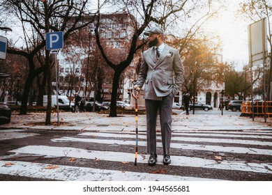 A black dapper bearded senior entrepreneur in an elegant custom-made costume, cap, with a fancy colorful walking stick is looking aside while crossing a road at a pedestrian crossing on an autumn day