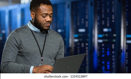 Black Data Center IT Technician Standing in Server Rack Corridor with a Laptop Computer. He is Visually Inspecting Working Server Cabinets.