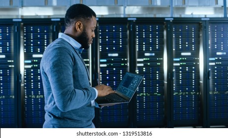 Black Data Center IT Technician Walking Through Server Rack Corridor with a Laptop Computer. He is Visually Inspecting Working Server Cabinets. - Powered by Shutterstock