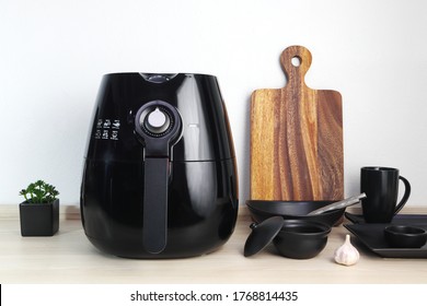 A Black Deep Fryer Or Oil Free Fryer Appliance, Mug, Dish And Wooden Tray Are On The Wooden Table In The Kitchen With A Small Plant In The Pot ( Air Fryer ) With Background Of White Cement Wall