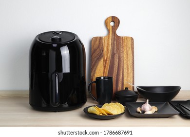 A Black Deep Fryer Or Oil Free Fryer Appliance, Mug, Dish And Wooden Tray Are On The Wooden Table In The Kitchen With A Small Plant In The Pot ( Air Fryer )