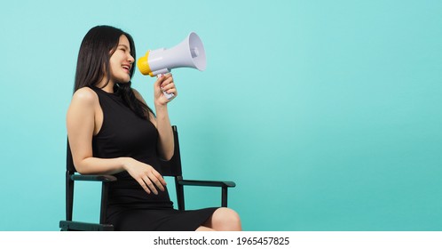 Black director chair and Asian woman is sit on it.She hold megaphone on mint color or Tiffany Blue background.
