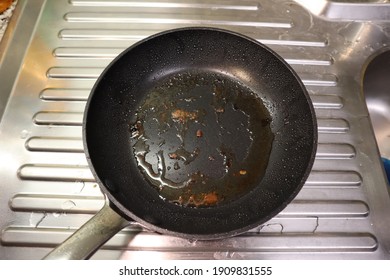 A black, dirty non stick frying pan with grease splatter and food residue. A stainless steel drain can be seen in the background. - Shutterstock ID 1909831555
