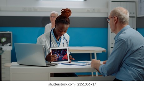 Black Doctor Holding Virus Animation On Tablet Showing Disease Cells To Elder Patient At Desk. African American Medic Explaining Coronavirus Bacteria Using Modern Technology On Device.