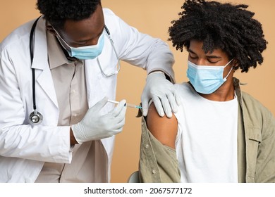 Black Doctor Inserting Syringe To Male Patient Arm, Making Vaccine Shot Against Coronavirus, Young African American Man Wearing Medical Mask Getting Vaccinated In Clinic, Beige Studio Background