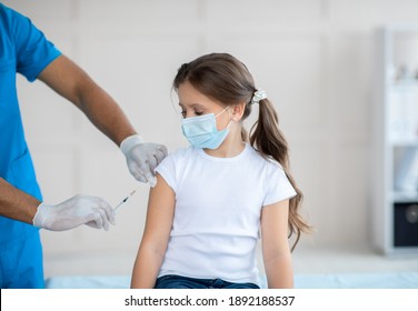 Black Doctor Making Coronavirus Vaccine Injection To Little Girl In Protective Mask At Hospital. Child Being Immunized Against Covid-19 At Health Centre. Global Vaccination Campaign Concept