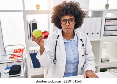 Black Doctor Woman With Curly Hair Holding Green Apple Thinking Attitude And Sober Expression Looking Self Confident 