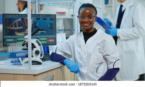 Black Doctor Woman Smiling At Camera Sitting In Scientific Modern Equipped Lab. Multiethnic Team Examining Virus Evolution Using High Tech, Chemistry Tools For Scientific Research, Vaccine Development