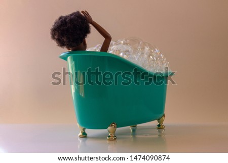 Black doll in miniature bathtub with foam, green plastic, golden feet. Soft background.