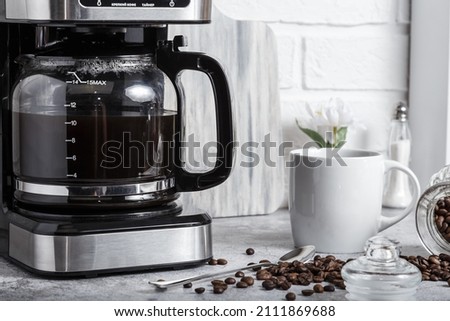 A black drip electric coffee machine with a glass teapot brews a morning drink. Household appliances, a white cup and a jar of beans on the kitchen table on the countertop at home