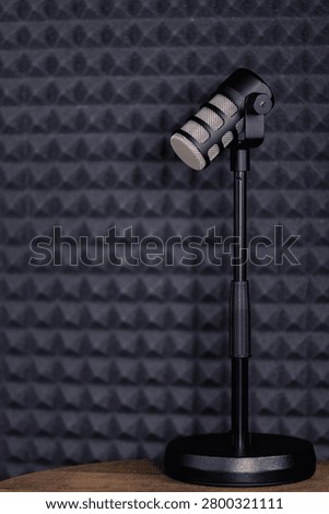 Black dynamic microphone mounted on an adjustable desktop stand, placed on a wooden surface against a dark grey acoustic foam panel background, ideal for podcasting, voiceover, or streaming. High quality, with copy space.