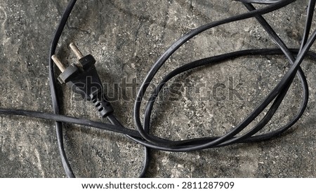 A black electrical plug with two prongs lies on a rough, textured surface, with its cord loosely coiled.