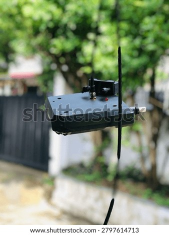 Black electronic device with a small red LED light and antenna connector hanging outdoors against a blurred green garden background.