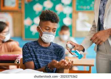 Black elementary student disinfecting hands with help of his teacher while wearing protective face mask in the classroom at school during coronavirus pandemic.  - Powered by Shutterstock