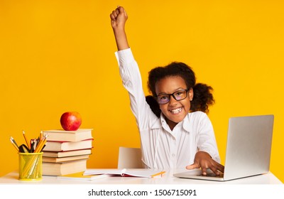 Black Elementary Student Girl Raising Hand Sitting At Laptop Doing Homework Over Yellow Background. Studio Shot