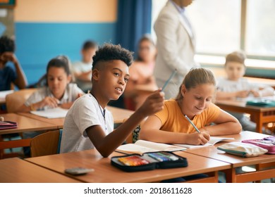 Black Elementary Student Talking To His Classmate And Pointing At Something On Blackboard During A Class At School. 