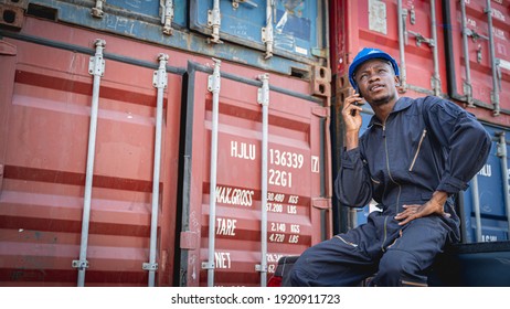 Black Employee In Blue Jumpsuit Sitting On The Back Of The Truck And Making Phone Call. Worker Taking A Break At Shipping Yard.