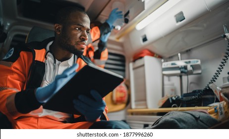 Black EMS Professional Paramedic Using Tablet Computer To Fill A Questionnaire For The Injured Patient On The Way To Hospital. Emergency Care Assistant Comforting The Patient In An Ambulance.