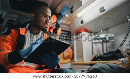 Black EMS Professional Paramedic Using Tablet Computer to Fill a Questionnaire for the Injured Patient on the Way to Hospital. Emergency Care Assistant Comforting the Patient in an Ambulance.