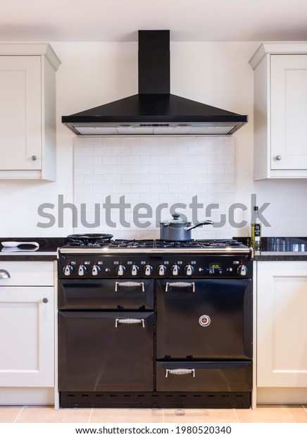 Black enamel oven range cooker with chimney hood\
in a modern kitchen with shaker style painted wood units in neutral\
off white. UK farmhouse\
kitchens