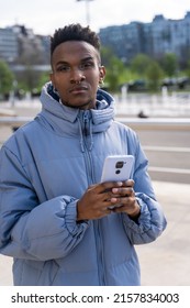 A black ethnic man with a phone and a blue jacket in the city, looking at social networks or digital press