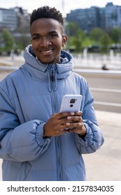 A black ethnic man with a phone and a blue jacket in the city, looking at social networks or digital press