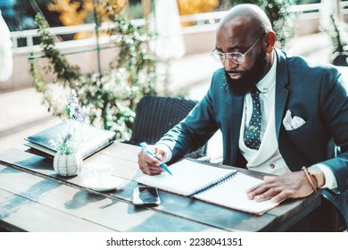 A Black executive man with a beard sitting on an outdoor terrace, dressed in a dark suit, polka-dot necktie, and a white vest, reviews paperwork, has a pen in his hand, and with his phone next to him - Shutterstock ID 2238041351
