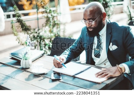 A Black executive man with a beard sitting on an outdoor terrace, dressed in a dark suit, polka-dot necktie, and a white vest, reviews paperwork, has a pen in his hand, and with his phone next to him