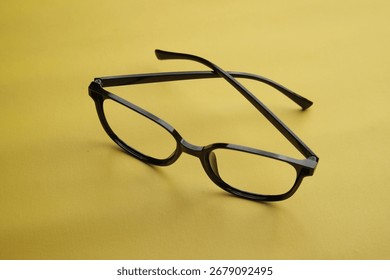 Black eyeglasses with a rectangular frame placed on a yellow surface. The frame is made of plastic material