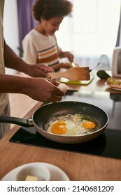 Black Family Cooking Breakfast At Home Kitchen. Focus On Foreground Of Father Frying Eggs On Pan With Background Of Blur Son Smearing Kiwi On Bread Piece. Relationship. Fatherhood. Domestic Lifestyle