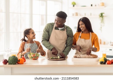 Black Family Cooking Together Making Salad Cutting Vegetables And Bread Standing In Modern Kitchen Indoors. Parents And Little Daughter Preparing Dinner. Healthy Food And Nutrition