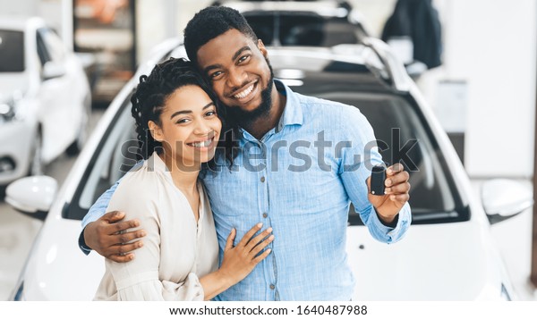 Black Family Couple Showing New Car Key Smiling\
To Camera In Dealership Showroom. Happy Spouses Hugging Near Their\
Auto. Panorama