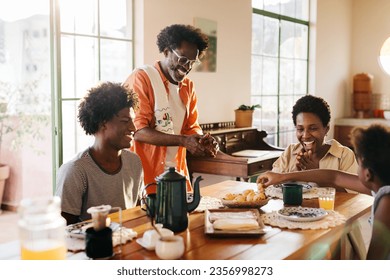 Black family enjoys a Brazilian breakfast in the kitchen at home. Father serves cheese bread rolls to his wife and kids, and everyone is smiling and bonding over the delicious, traditional meal. - Powered by Shutterstock