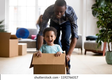 Black family in living room have a fun spend time at new home. African adorable playful laughing boy sitting at cardboard box, father rolling him playing together. New property and relocation concept