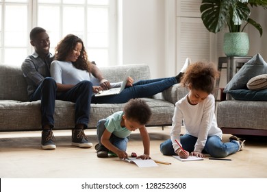 Black Family Relaxing At Modern Home, African American Parents Resting On Sofa With Laptop While Mixed Race Kids Playing Together Drawing On Warm Floor, Children Have Fun With Mom Dad In Living Room