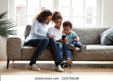 Black Family Sitting On Couch Have A Fun At Home. African Mother Spend Free Time With Daughter And Son Using Mobile Phone Taking Selfie Photo Playing New Game. Happy Family Together On Weekend Concept